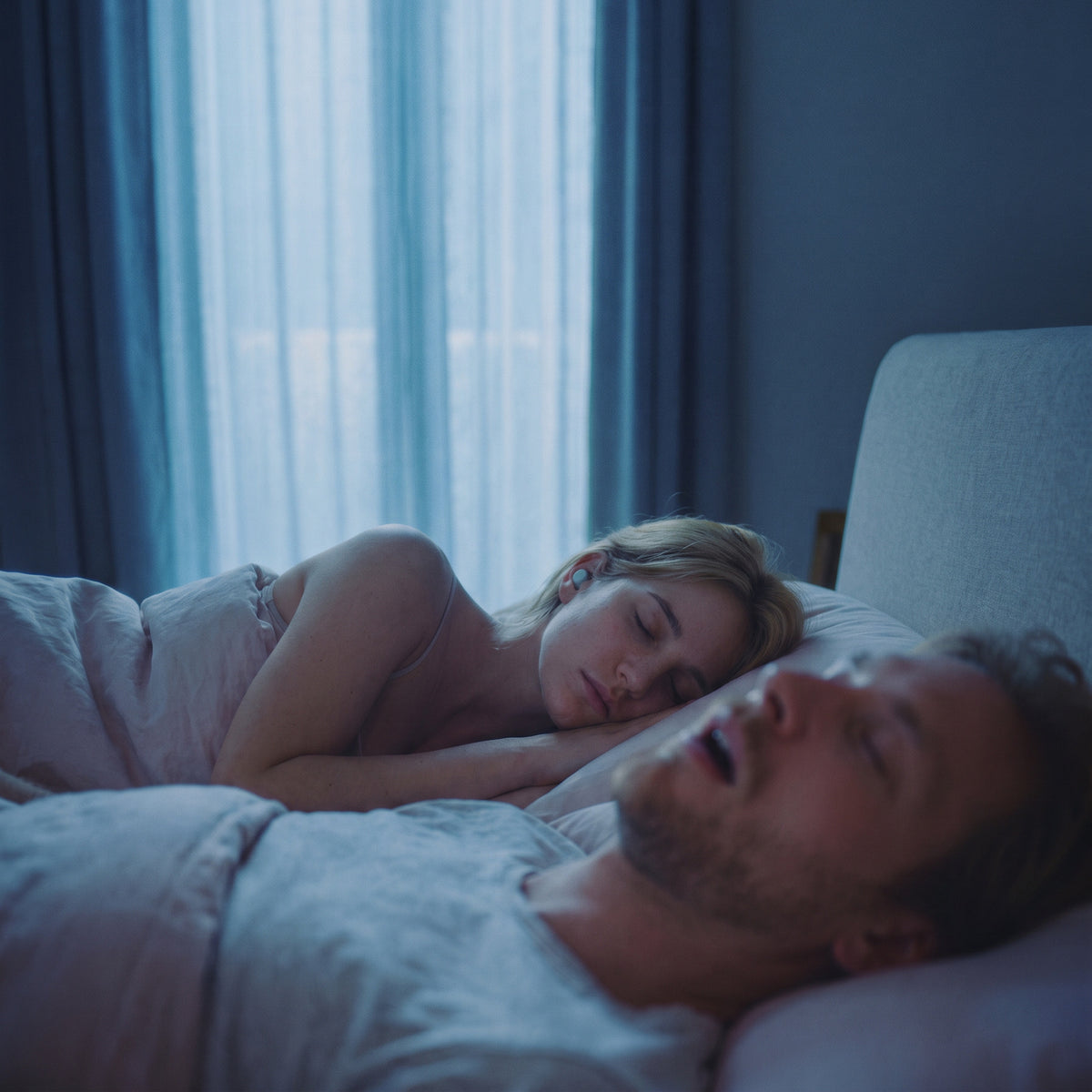Nachts im Schlafzimmer: Vorn schläft ein Mann mit offenem Mund und schnarcht, dahinter schläft eine blonde Frau ruhig, im Ohr ein hellgrauer Loume Dream Dots Kopfhörer.